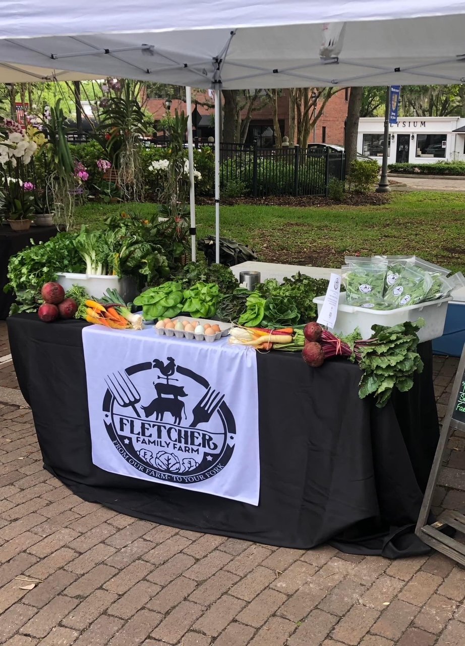 Fresh produce display at Fletcher Family Farm booth, featuring a variety of leafy greens, colorful carrots, beets, and farm-fresh eggs, set up under a canopy at a farmers' market.